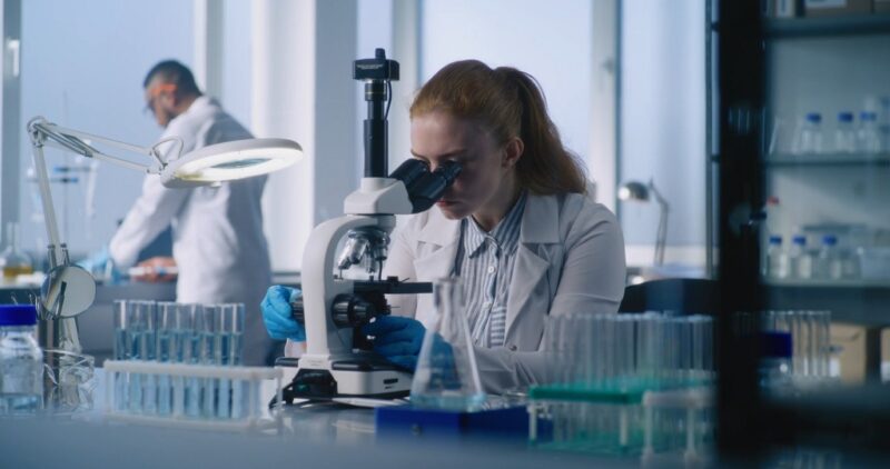 Female scientist analyzing samples through a microscope in a modern laboratory, representing evidence-based scientific research and transparency
